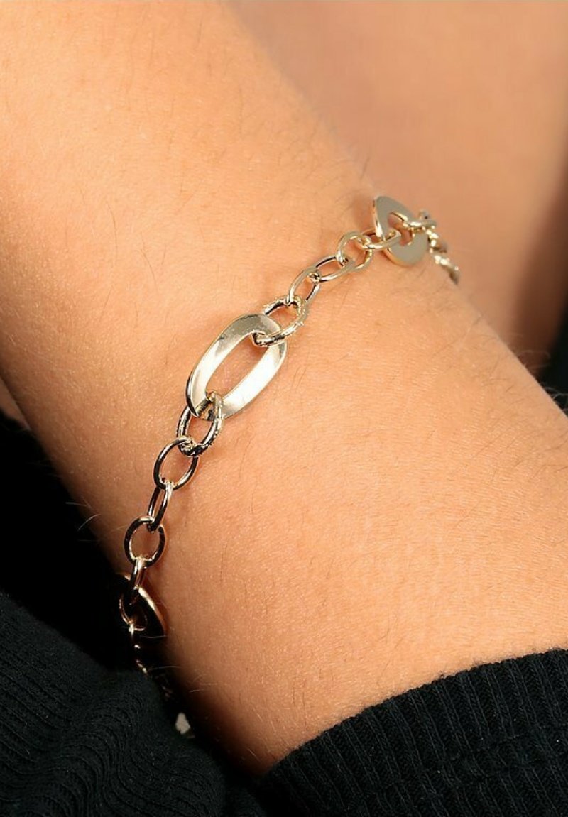 Das goldene Kettenarmband verfügt über ovale und kreisförmige Glieder mit einer glatten Oberfläche. Das Armband wird am Handgelenk getragen und steht im Kontrast zum Hautton.