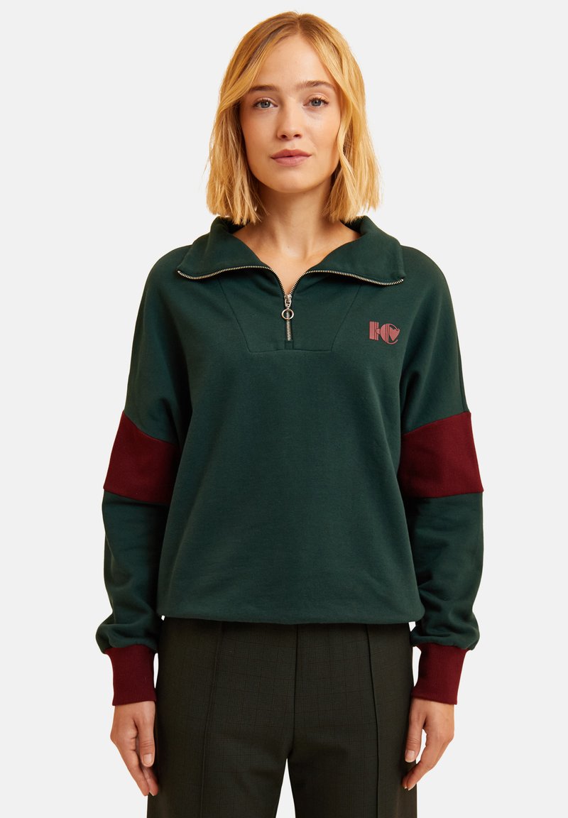 Sudadera verde con cuello de cremallera, con acentos en color marrón en las mangas y un logo en el pecho. Tejido suave con un corte relajado.