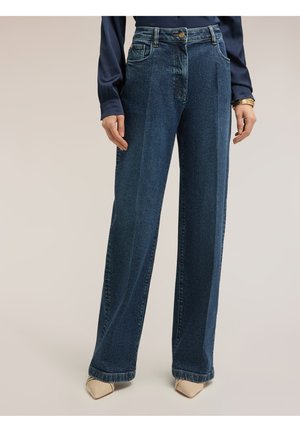 Straight-leg dunkelblaue Jeans, getragen mit beigen spitzen Schuhen und einem marineblauen Langarmshirt auf neutralem Hintergrund.