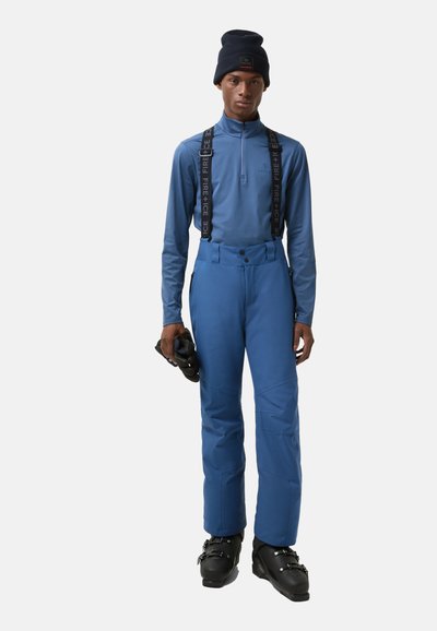 Niebieski strój narciarski z wysokim kołnierzem i szelkami, o gładkiej fakturze. Zawiera buty narciarskie trzymane w jednej ręce.