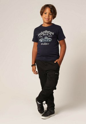 T-shirt graphique bleu marine avec impression de camion blanc et texte, associé à un pantalon cargo noir avec poches latérales et des baskets noires.