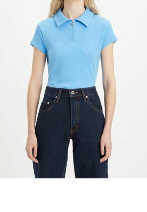 Frau trägt ein hellblaues, kurzärmeliges, geripptes Oberteil mit Reißverschluss am Kragen und hoch taillierte dunkelblaue Jeans, steht vor weißem Hintergrund.