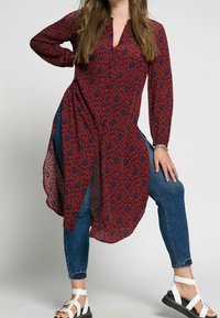 Tunique rouge avec un motif abstrait noir et bleu, fabriquée en tissu léger, dotée de manches longues et d'un ourlet asymétrique. Portée avec un jean bleu.