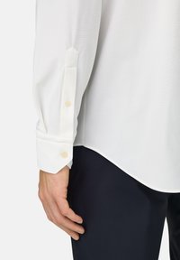 Weißes Hemd mit langen Ärmeln und geknöpften Manschetten, versehen mit einer strukturierten Stoffoberfläche und spitzen Manschetten, kombiniert mit dunkelblauen Hosen.