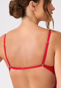 Czerwona koronkowa braletka z cienkimi ramiączkami i złotymi elementami, pokazująca gładkie plecy z kwiatowym wzorem wzdłuż krawędzi.