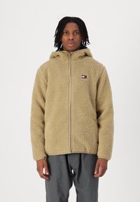 Beige Fleecejacke mit Kapuze, ausgestattet mit einem Frontreißverschluss und einem kleinen Logopfpatch. Strukturiertes Material und lässige Passform. Wird mit grauen Hosen getragen.
