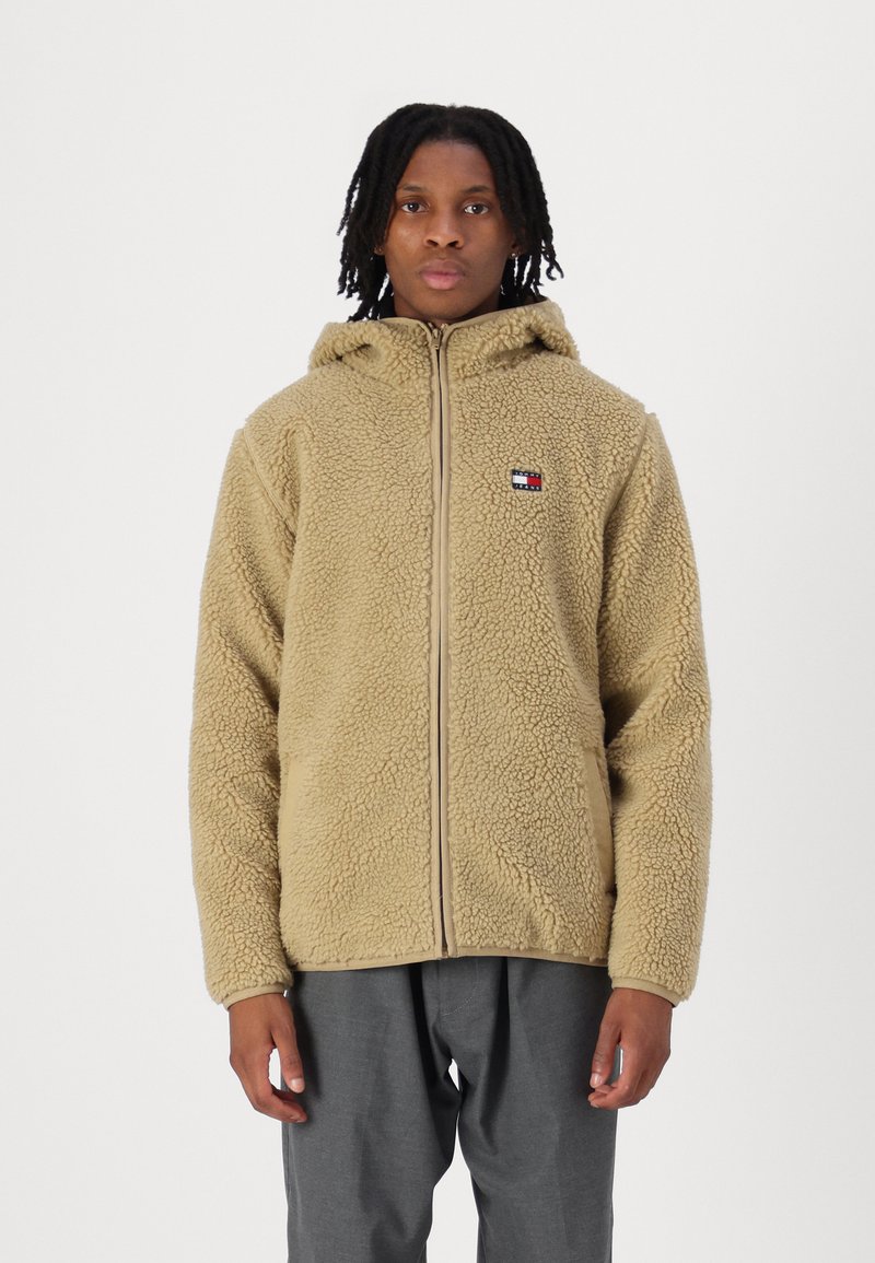 Beige Fleecejacke mit Kapuze, ausgestattet mit einem Frontreißverschluss und einem kleinen Logopfpatch. Strukturiertes Material und lässige Passform. Wird mit grauen Hosen getragen.