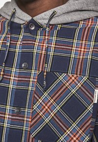 Koszula w kratę w kolorach granatowym, pomarańczowym, żółtym i szarym; posiada szary kaptur, zapięcie na guziki oraz kieszeń naszywaną z wzorem w kratę.