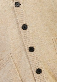 Beige kofta av mjukt tyg med ribbad halsringning, försedd med sex svarta knappar och två framfickor.