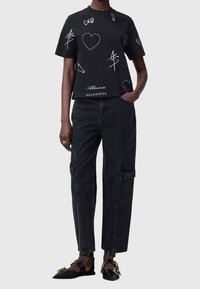 Mustat lyhyet t-paidat, joissa on valkoisia doodle-tyylisiä grafiikoita ja tekstejä, yhdistettyinä löysiin mustiin cargo-housuihin ja mustiin sandaaleihin.