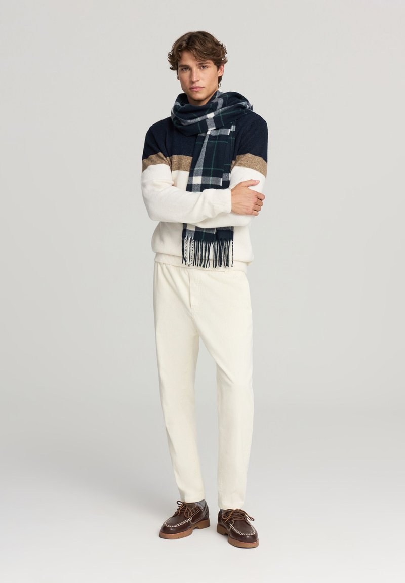 Giovane uomo in piedi con le braccia incrociate, indossa un maglione color-block, pantaloni bianchi, scarpe marroni e una sciarpa a quadri scura su uno sfondo neutro.