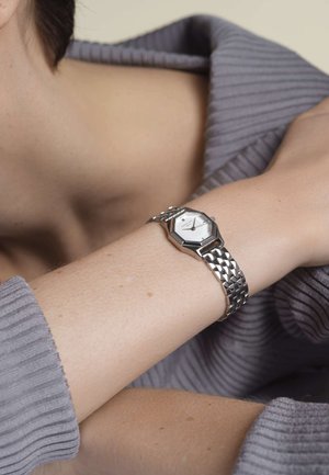 Montre octogonale argentée avec un cadran blanc, bracelet en maillons métalliques et surface texturée, portée sur un bras pâle contre un pull gris à côtes.