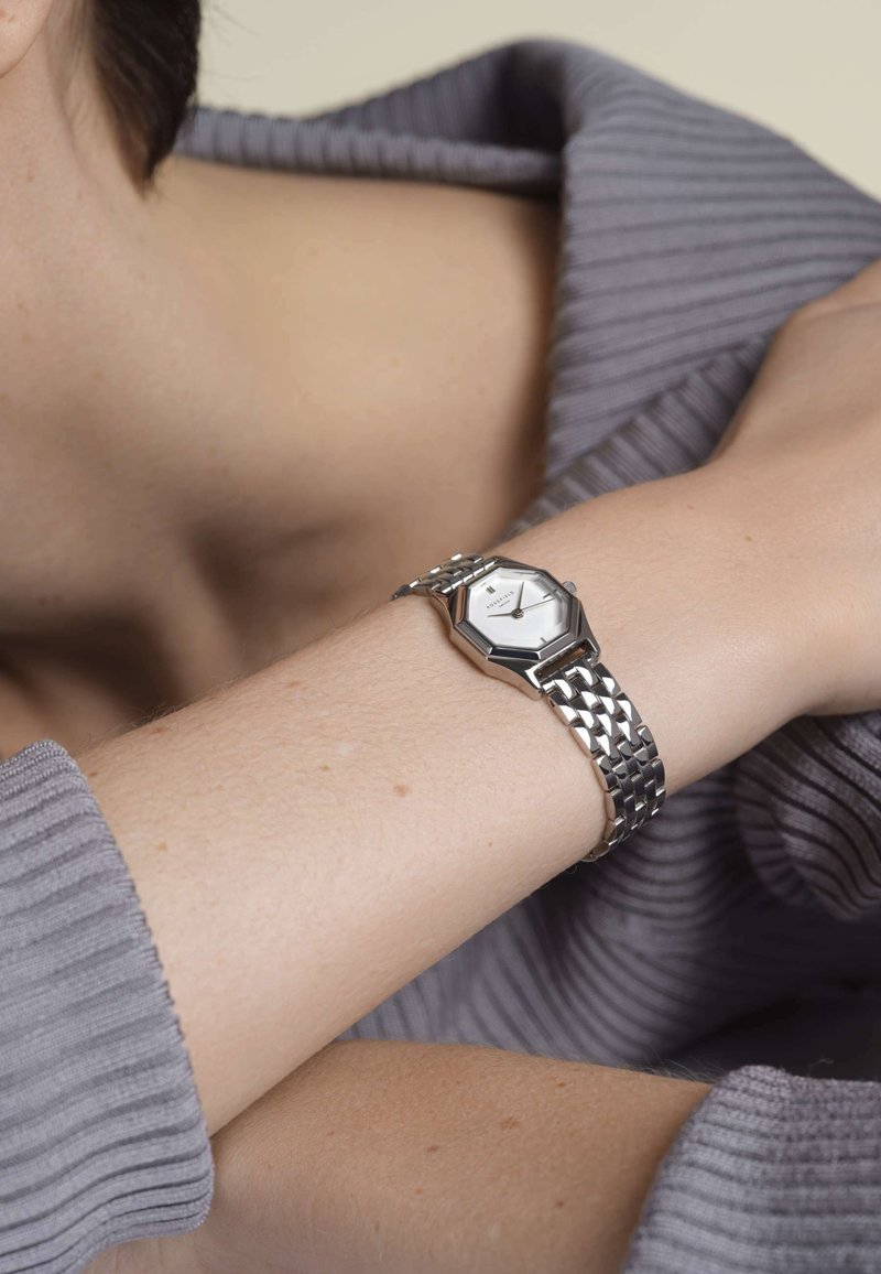 Srebrni osmerokutni sat s bijelim licem, metalnom narukvicom i teksturiranom površinom, nosi se na svijetloj ruci protiv sivog rebrastog pulovera.