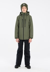 Giacca verde oliva con tasche sul petto con zip, abbinata a pantaloni da neve neri e stivaletti neri, con un berretto di lana verde.