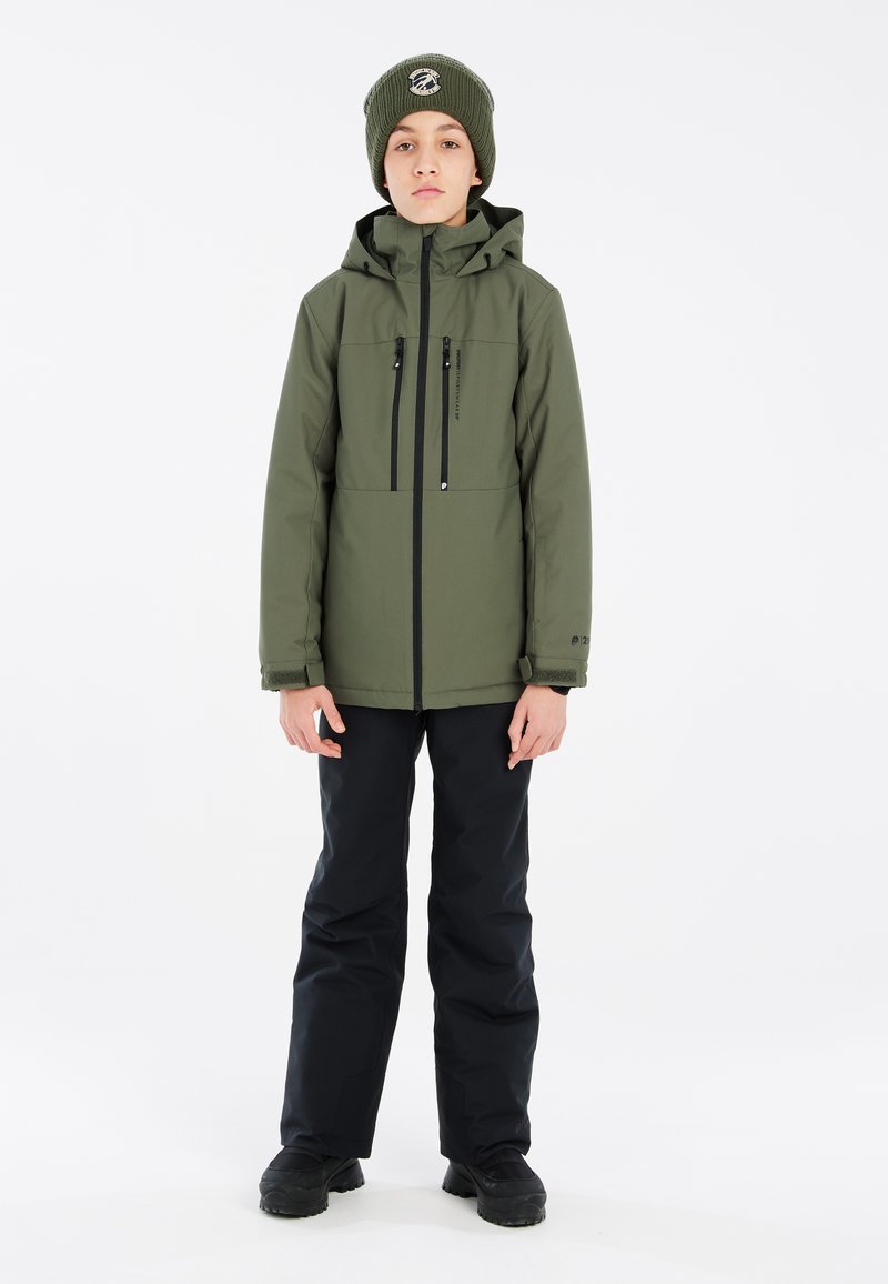 Giacca verde oliva con tasche sul petto con zip, abbinata a pantaloni da neve neri e stivaletti neri, con un berretto di lana verde.