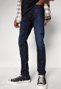 Jeans en denim bleu foncé à coupe slim, agrémentés de coutures contrastées. Portés avec des baskets noires à semelles blanches et lacets noirs.