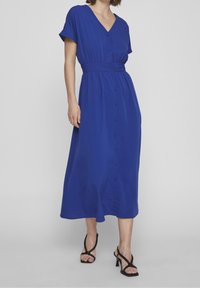 Modrá šaty s krátkým rukávem a véčkovým výstřihem, se staženým pasem a předním zapínáním na knoflíky. Mají splývavou sukni a hladkou texturu látky.