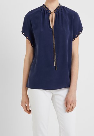 Femme portant un chemisier bleu marine à manches courtes avec des bords festonnés en œillet et un lien chaîne dorée, assorti à un pantalon blanc.