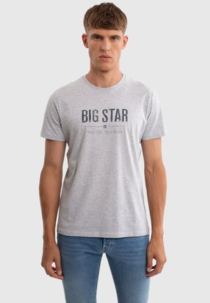 Young man wearing a light grey "BIG STAR" logo t-shirt and blue jeans, standing against a plain light background.