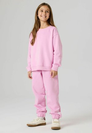 Młoda dziewczyna uśmiechająca się, ubrana w luźną różową bluzę i dopasowane różowe spodnie dresowe, w białych tenisówkach, stojąca na tle jednolitego, jasnego tła.