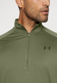 Zöld negyedcipzáras pulóver puha, textúrált anyagból. Cipzáras gallérral és egy diszkrét fekete Under Armour logóval a mellen.