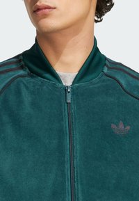 Giovane che indossa una giacca a zip in velluto teal con colletto a coste e logo Adidas ricamato sul lato sinistro del petto.