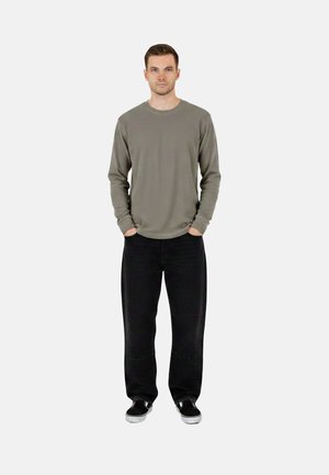 Uomo in piedi con le mani in tasca, che indossa una maglia grigia a maniche lunghe, pantaloni neri e scarpe slip-on nere su sfondo bianco semplice.