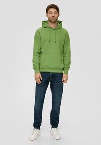 Sudadera verde de algodón con bolsillo frontal, cordones, puños elásticos, combinada con jeans de un azul oscuro y zapatillas blancas. Estilo sencillo y casual.