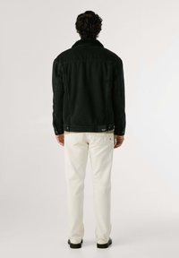Chaqueta de mezclilla negra con un cuello grueso y texturizado, corte relajado y costuras prominentes, llevada sobre pantalones blancos, mostrando un diseño minimalista.