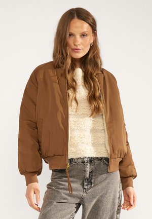 Woman with long wavy hair wearing a brown bomber jacket, cream textured sweater, and high-waisted gray jeans, standing against a plain background.