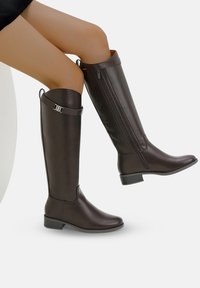 Bottes en cuir marron montantes avec une finition lisse, dotées d'une fermeture éclair sur le côté, d'accents de sangle et d'un petit talon carré.