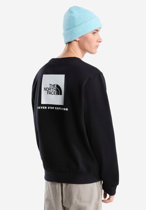 Jeune personne portant un sweat-shirt noir avec le logo The North Face et le texte "Never Stop Exploring" dans le dos, ainsi qu'un bonnet tricoté bleu clair.