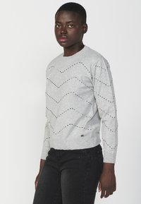 Maglione grigio lavorato a maglia con motivo a zigzag e piccoli fori, maniche e orlo a coste, abbinato a jeans scuri.