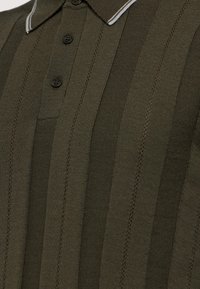 Polo tričko s olivově zelenými pruhy vyrobené z pleteného materiálu, s klasickým límcem, třemi černými knoflíky a jemným bílým akcentem v límci.