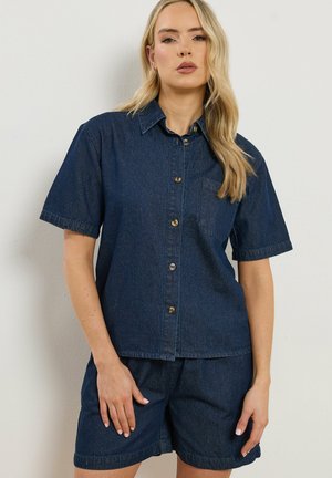 Vrouw draagt een donkerblauw denim overhemd met korte mouwen en bijpassende korte broek, staand tegen een eenvoudige lichte muur.