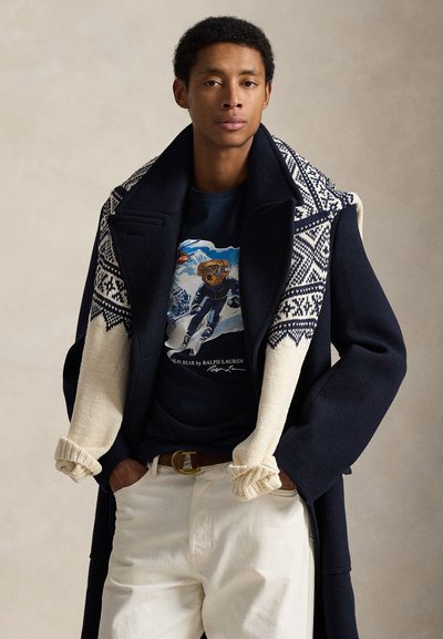 Navy wool coat with knit white sleeves featuring a geometric pattern. Paired with a dark T-shirt and cream-colored pants, accented by a belt.