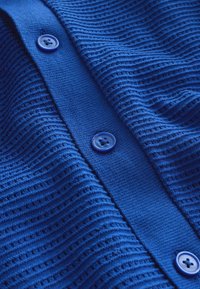 Cardigan bleu en maille avec nervures horizontales texturées, comportant trois boutons ronds bleus visibles sur le devant au centre.