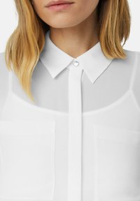 Witte doorzichtige blouse met een klassieke kraag, een knoopsluiting aan de voorkant en twee opgestikte zakken. De stof lijkt lichtgewicht en glad te zijn.