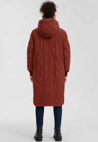 Langer, gesteppten, kapuzenmantel in rostfarbener Farbe mit Diamantmuster, ausgestattet mit gerippten Bündchen und einem geraden Saum. Kombiniert mit dunklen Jeans und Stiefeln.