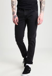 Jeans de mezclilla negra con un corte ajustado, textura deslavada, diseño de cinco bolsillos y detalles de costura. Llevados con zapatillas negras y puños enrollados.