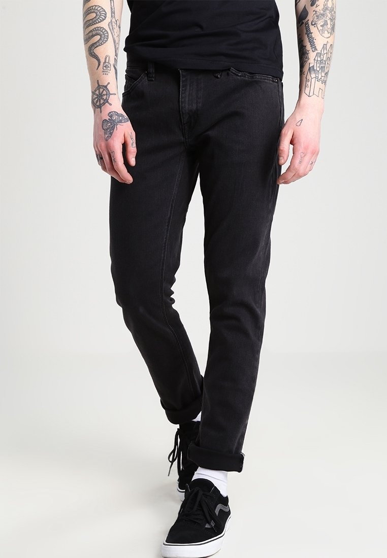 Jeans de mezclilla negra con un corte ajustado, textura deslavada, diseño de cinco bolsillos y detalles de costura. Llevados con zapatillas negras y puños enrollados.