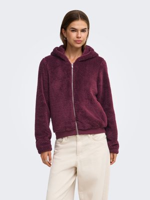 Femme portant une veste en peluche violette zippée à capuche avec un pantalon couleur crème, debout devant un fond clair uni.
