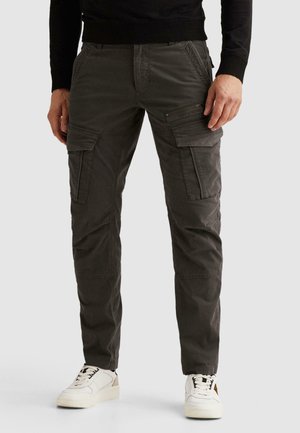 Mann trägt dunkelgraue Cargohose mit mehreren Taschen und weiße Turnschuhe vor einem einfarbigen Hintergrund.