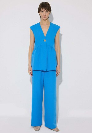 Femme portant un haut bleu vif sans manches à col en V avec un seul bouton doré et un pantalon large assorti, debout contre un mur clair.