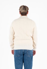 Cremefarbener Strickpullover mit gerippter Struktur und Kragen, ausgestattet mit langen Ärmeln und einem lässigen Schnitt, kombiniert mit blauen Jeans.
