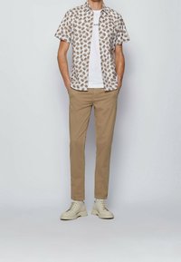 Bílé tričko s krátkým rukávem a hnědým květinovým vzorem přes bílé tričko, doplněné béžov�ými kalhotami a světle zbarvenými robustními botami.