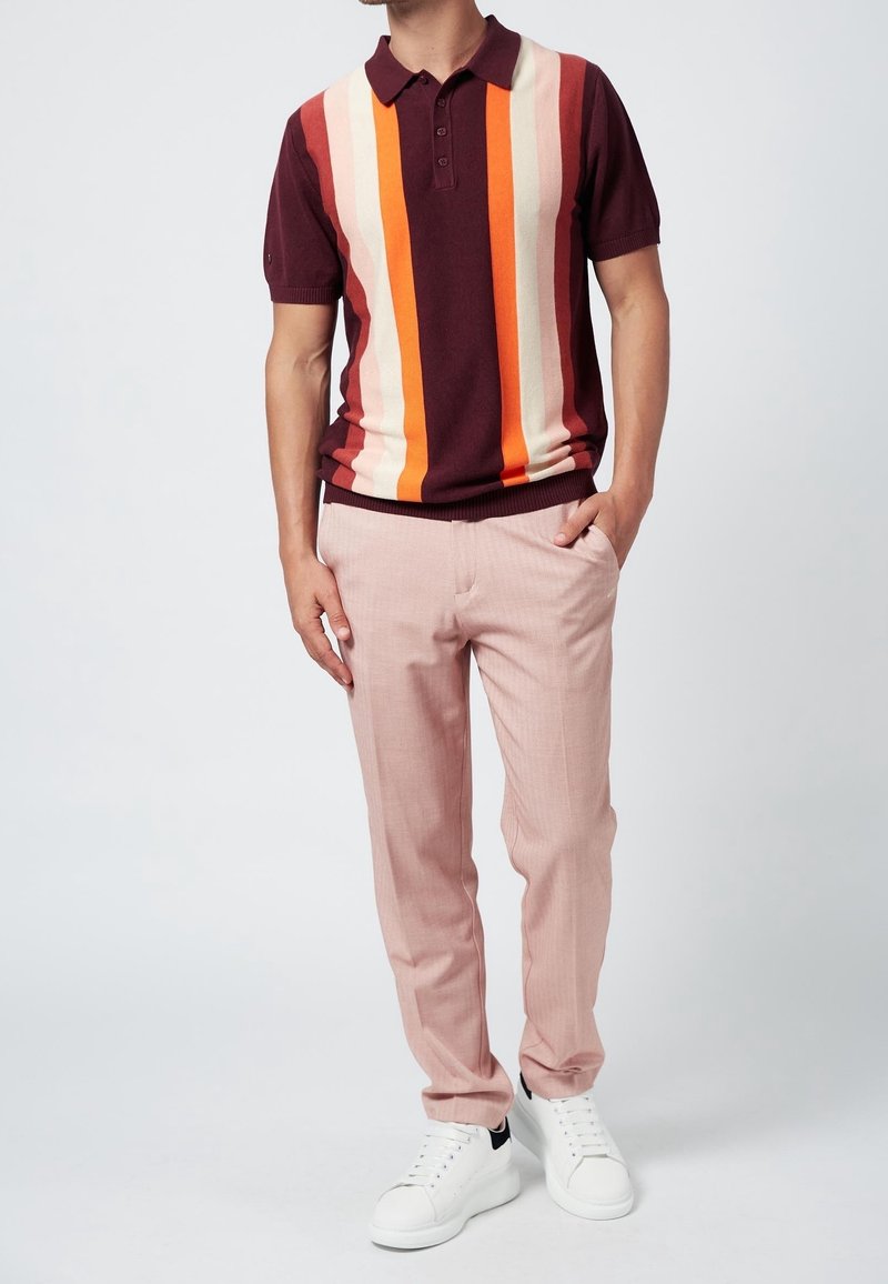 Mies, ktorý má na sebe polo tričko s krátkym rukávom s vertikálnymi pruhmi v marhuľovej, oranžovej a béžovej farbe, ružové nohavice a biele tenisky.