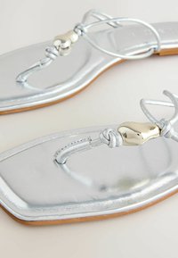 Sandales plates métalliques argentées avec lanières en forme de tongs et embellissements en forme de cœur doré, présentées en gros plan sur un fond clair.