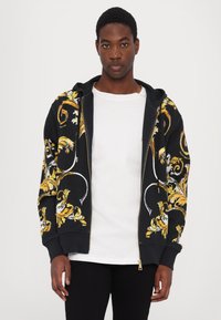 Felpa nera con zip, decorata con motivi floreali gialli e bianchi, caratterizzata da un cappuccio con coulisse e polsini a coste. Indossata sopra una maglietta bianca.