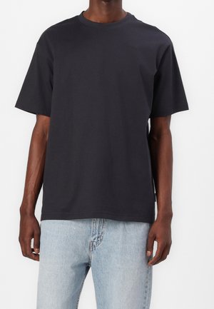 Mand iført en simpel sort T-shirt med korte ærmer og lyseblå denimjeans står mod en hvid baggrund.
